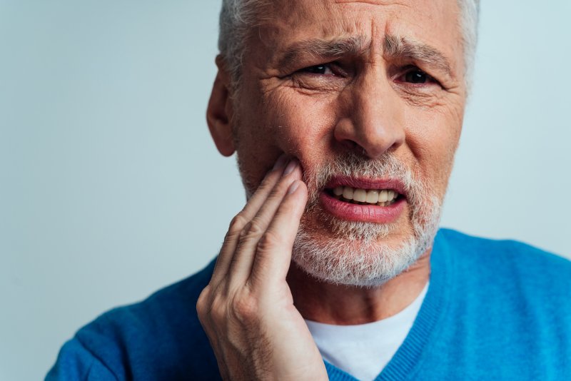 Older man wearing blue shirt holding right hand to cheek and grimacing in pain
