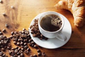 Cup of coffee on white saucer surrounded by coffee beans and croissant.